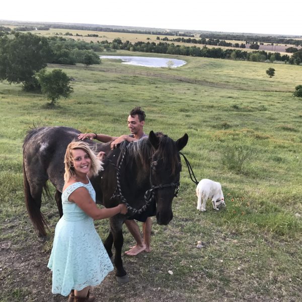 couple posing with one of our quarter horses, Moonshine.  Woman in blue floral dress smiling on the bluff top of our Texas dude ranch while her partner grins and prepares to mount up bareback.  White Akbash dog, Cecelia, stands by waiting for something fun to do in Austin 