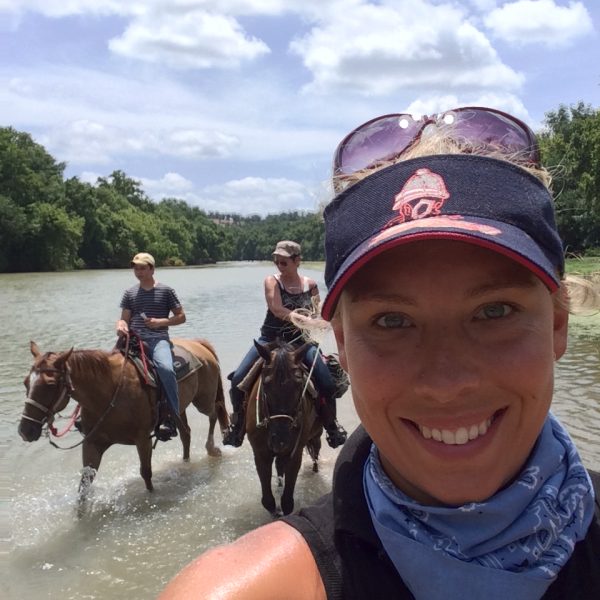 Natural horsemanship trainer MacCoy poses with her students in the background, enjoying the river of McKinney Roughs in Bastrop state park on a horseback riding adventure