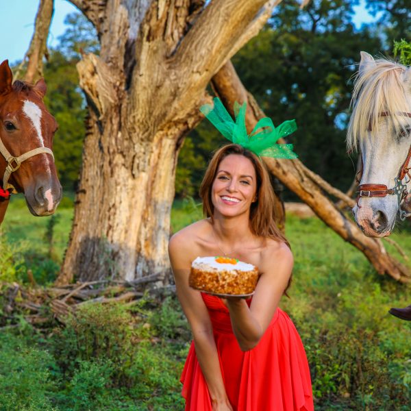 Beautiful horse model with orange dress and green head piece reminiscent of a carrot teases hungry horses in the background, in the middle of green fields at a Round Rock horseback riding lesson stables in Texas