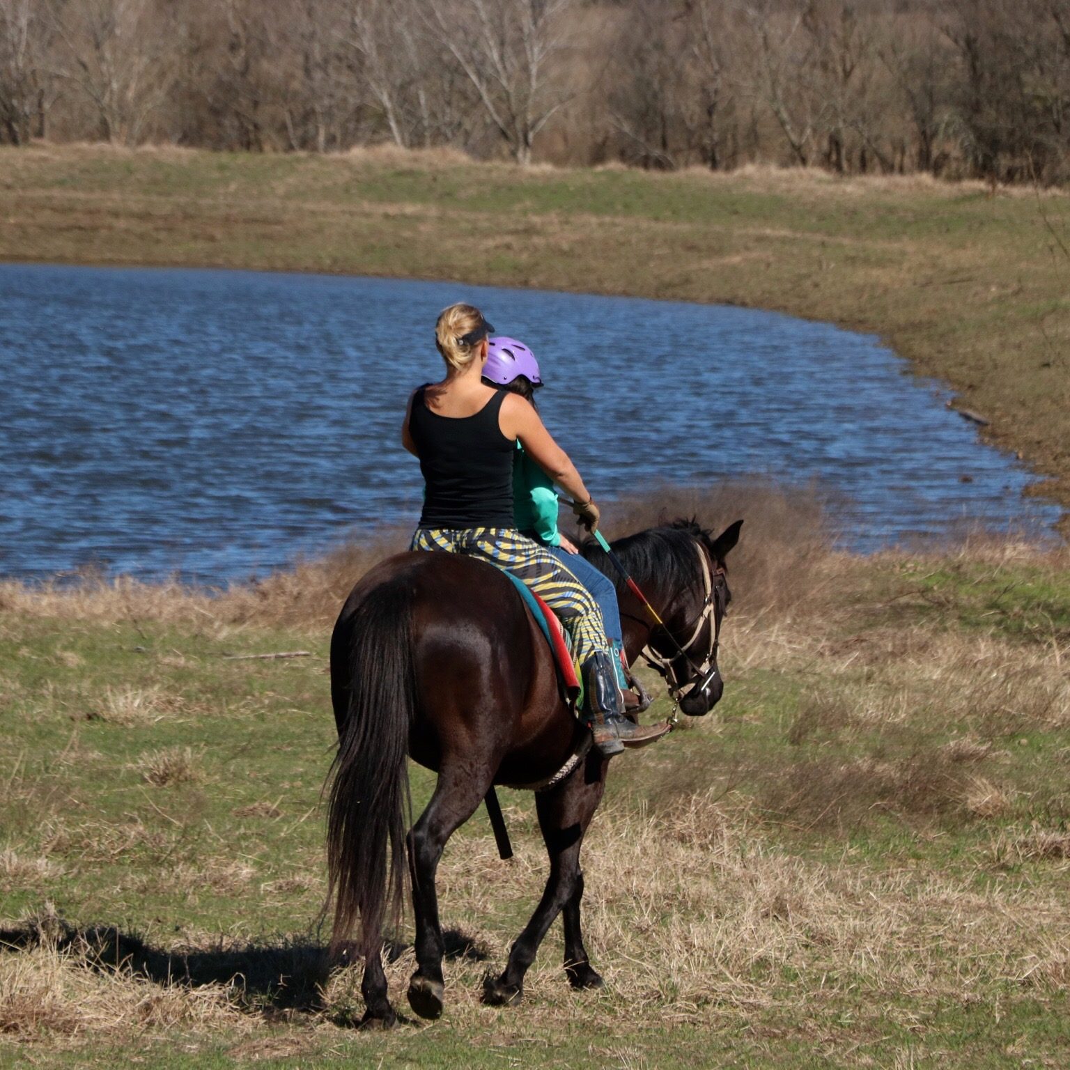 horse trainer MacCoy rides black horse in front of large pond with equestrian riding student
