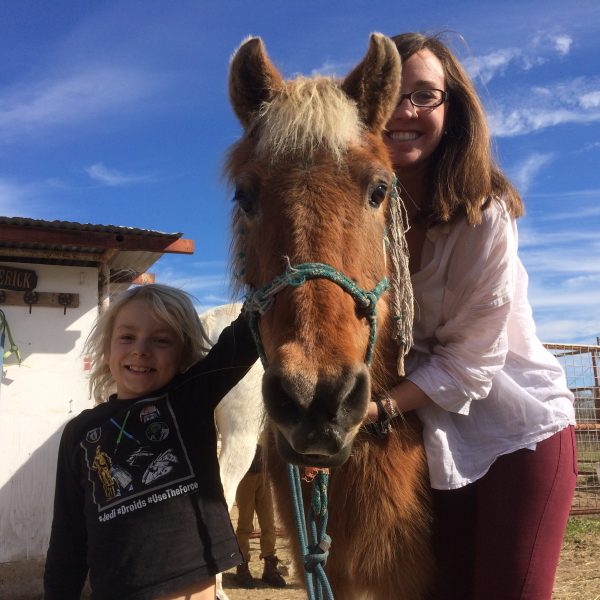 Blonde boy in long sleeve shirt stands next to his beloved blonde pony named Texas Glory, standing with horseback riding instructor Kathryn Hetzendorfer