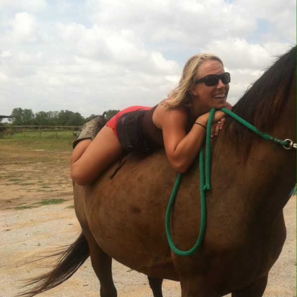 blonde woman in brown tank top and red shorts sporting cowboy boots lying on big bay gelding bareback, holding green lead line with sunglasses and a big smile