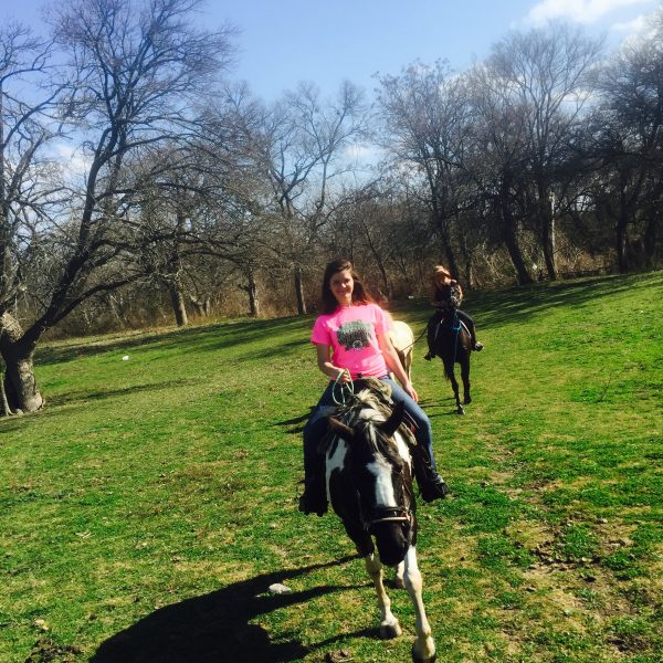 Forward shot by equine photographer who is mounted, but not pictured, of black and white paint horse with white blaze in green pastures in Hutto, riding with student in bright pink shirt and horse trainer bringing up the rear 
