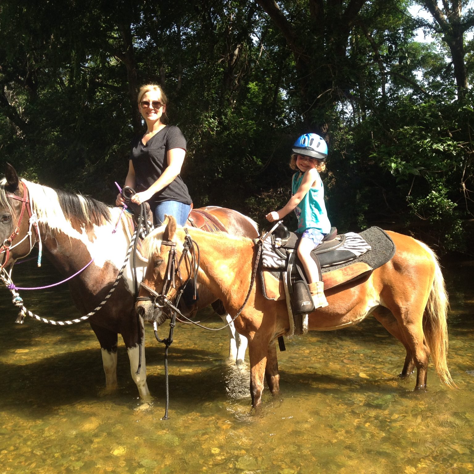 Smiling little girl on cute flaxen mane pony standing in the water with mother, mounted on gorgeous black and white paint mare for sale in San Marcos, Texas