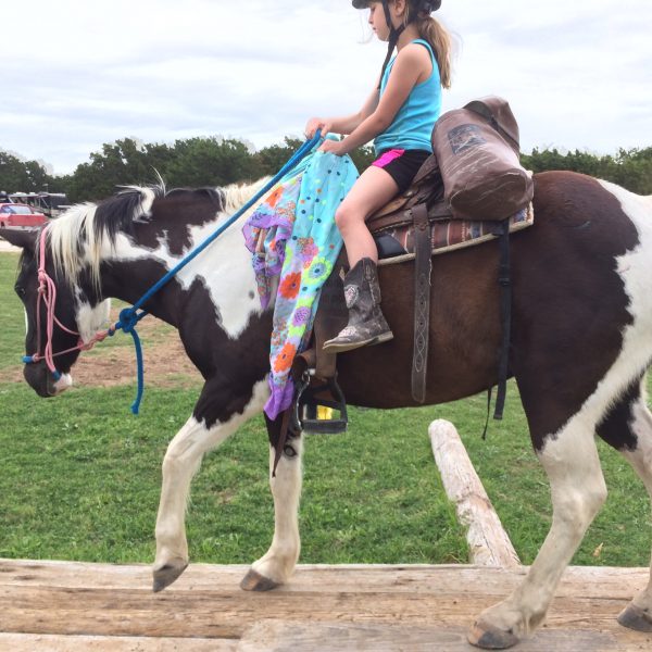Black and white paint mare walks on horseback riding obstacle with youth play day rider, eaight years old, pictured in shorts and blue tank top